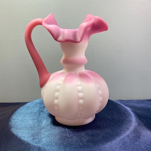 May include: A white and pink glass pitcher with a ruffled top and a textured body. The pitcher has a pink handle.
