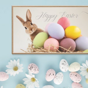 Puede incluir: Una impresi&oacute;n enmarcada con un conejo marr&oacute;n asom&aacute;ndose de un nido de heno con huevos de Pascua coloridos. La impresi&oacute;n dice "Feliz Pascua" en una fuente cursiva.