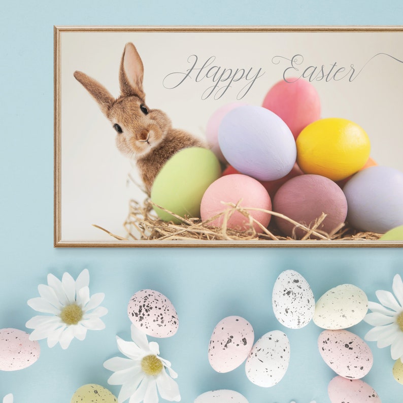 Puede incluir: Una impresi&oacute;n enmarcada con un conejo marr&oacute;n asom&aacute;ndose de un nido de heno con huevos de Pascua coloridos. La impresi&oacute;n dice "Feliz Pascua" en una fuente cursiva.