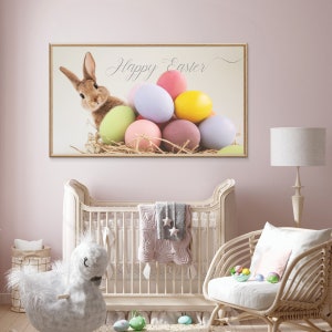 Puede incluir: Una impresi&oacute;n enmarcada con un conejo marr&oacute;n asom&aacute;ndose desde un nido de huevos de Pascua de colores pastel. La impresi&oacute;n tiene el texto "Happy Easter" en una fuente cursiva.