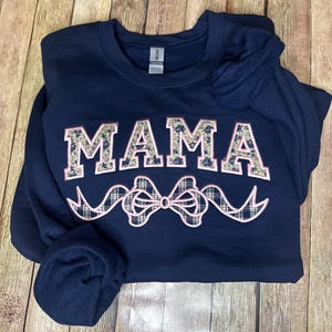 May include: A navy blue sweatshirt with the word "MAMA" in large, white letters with a camouflage pattern. The letters are outlined in pink. Below the word "MAMA" is a pink plaid bow with white ribbon tails.
