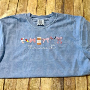 May include: Light blue t-shirt with embroidered pharmacy-themed designs, including pills, bows, a prescription bottle, and a mortar and pestle. The word "Pharmacist" is embroidered below the designs.