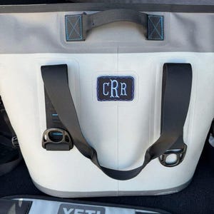 Könnte beinhalten: Eine weiß-graue Kühlbox mit schwarzen Trägern und dem Text "YETI". Die Kühlbox hat einen blau gestickten Aufnäher mit den Initialen "CRR". Die Kühlbox hat einen schwarzen Griff und einen grau-weißen Schultergurt.