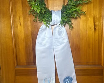 May include: A green wreath with a white satin ribbon. The ribbon has a light blue embroidered initial and a blue hydrangea flower. The wreath is hanging on a wooden door.