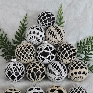 May include: A collection of 13 crocheted Christmas ornaments in black, white, and cream. The ornaments are arranged in a pyramid shape with a green sprig of cedar behind them.