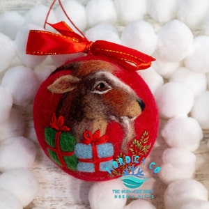 May include: Red felt Christmas ornament with a hand-felted goat head and a green and blue gift box. The ornament is decorated with gold thread and has a red ribbon attached. The ornament is labeled "Nordica Co. The Ornament Shop Nordic Gifts."