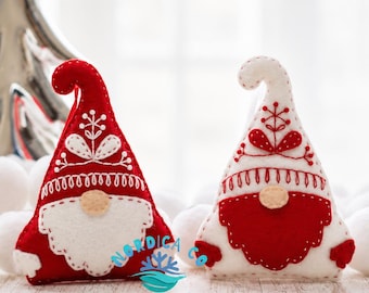 May include: Two felt gnome ornaments, one red with white embroidery and one white with red embroidery, each featuring a pointed hat, a round nose, and a beard. The ornaments are decorated with stitched floral patterns and include the text NORDICA CO.