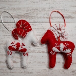 May include: Two felt Christmas ornaments shaped like reindeer with red and white details. The reindeer on the left is white with red antlers and a red floral design. The reindeer on the right is red with white antlers and a white floral design.