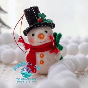 May include: A felt snowman ornament featuring a black top hat with green holly and a red berry, a bright red scarf, an orange carrot nose, and two orange buttons. The snowman has a red hanging string and the text NORDICA CO THE ORNAMENT SHOP NORDIC GIFTS.