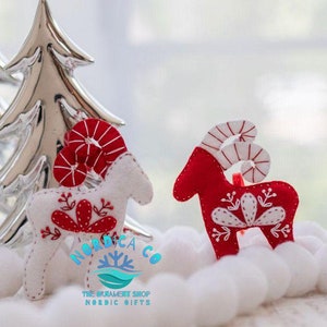 May include: Two felt Christmas ornaments shaped like reindeer with red and white candy cane horns. The reindeer are standing on a white surface. The reindeer on the left is white with red details and the reindeer on the right is red with white details. The ornaments are handmade and have a Scandinavian design.