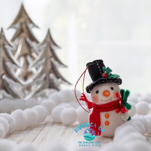 May include: A felt snowman ornament wearing a black top hat with green holly and a red berry accent, a bright red scarf, and orange buttons. The snowman has a carrot-shaped nose, rosy cheeks, and a red hanging string. Text reads NORDICA CO.