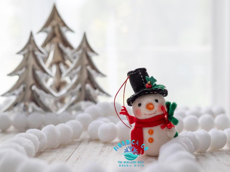 May include: A felt snowman ornament wearing a black top hat with green holly and a red berry accent, a bright red scarf, and orange buttons. The snowman has a carrot-shaped nose, rosy cheeks, and a red hanging string. Text reads NORDICA CO.