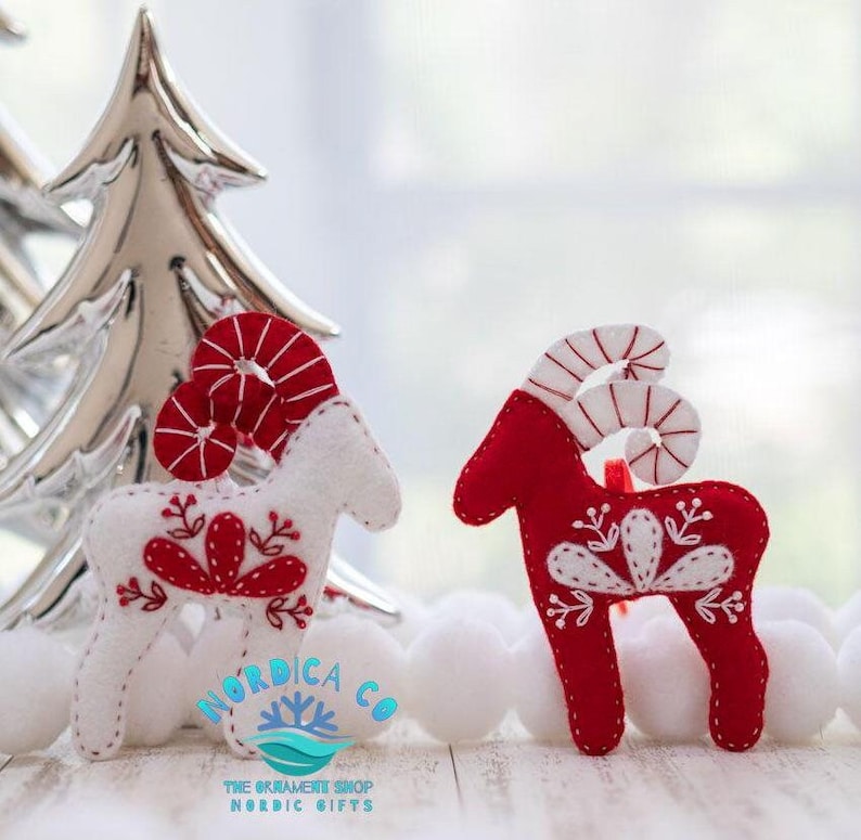 May include: Two felt Christmas ornaments shaped like reindeer with candy cane antlers. One reindeer is white with red embroidery and the other is red with white embroidery. The ornaments are on a white wooden surface with a silver Christmas tree in the background.
