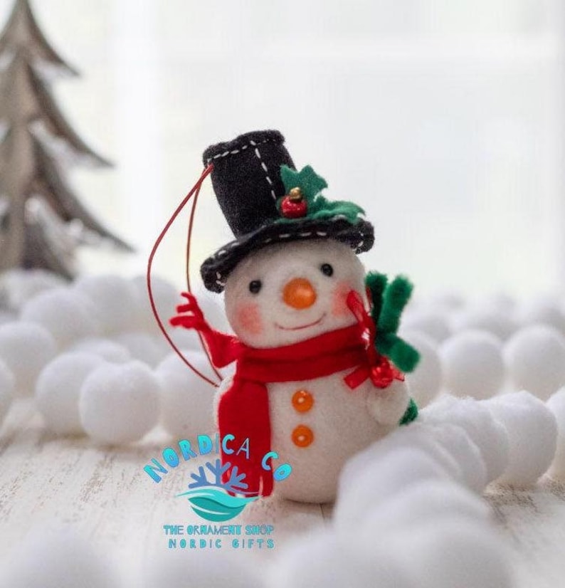 May include: A felt snowman ornament featuring a black top hat with green holly and a red berry, a bright red scarf, an orange carrot nose, and two orange buttons. The snowman has a red hanging string and the text NORDICA CO THE ORNAMENT SHOP NORDIC GIFTS.