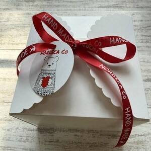 May include: A square white gift box with scalloped edges, tied with a red satin ribbon featuring the text "HAND MADE" and "NORDICA CO". A round white tag is attached to the ribbon, displaying a line-art illustration of a bear holding a red heart.