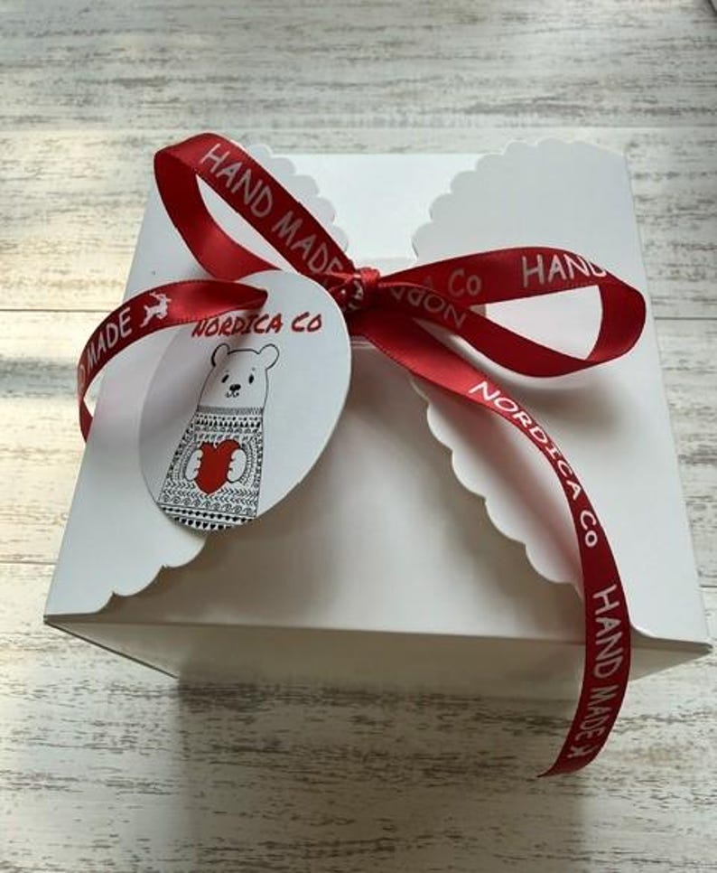 May include: A square white gift box with scalloped edges, tied with a red satin ribbon featuring the text "HAND MADE" and "NORDICA CO". A round white tag is attached to the ribbon, displaying a line-art illustration of a bear holding a red heart.