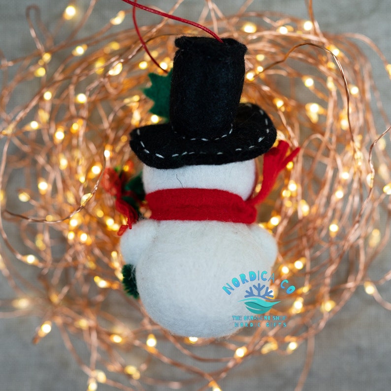 May include: A felt snowman ornament featuring a white rounded body, a black top hat with white stitching, and a bright red scarf. The snowman holds a small green branch and is suspended by a thin red string. The logo NORDICA.CO is visible.