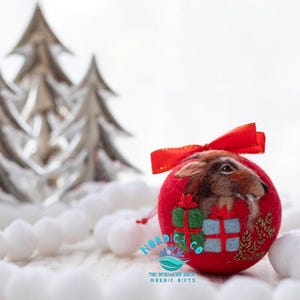 May include: A round red felt ornament featuring a needle-felted brown hare, two small gift boxes in green and blue, and gold embroidered foliage. The ornament is topped with a red ribbon bow and displays the text NORDICA CO THE ORNAMENT SHOP NORDIC GIFTS.