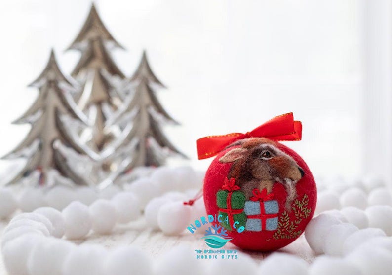 May include: A round red felt ornament featuring a needle-felted brown hare, two small gift boxes in green and blue, and gold embroidered foliage. The ornament is topped with a red ribbon bow and displays the text NORDICA CO THE ORNAMENT SHOP NORDIC GIFTS.