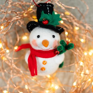 May include: A felt snowman ornament wearing a black top hat with green holly leaves and a red berry, a bright red scarf, and two orange buttons. The snowman has a carrot-shaped nose, pink cheeks, and holds a small green branch with red berries.