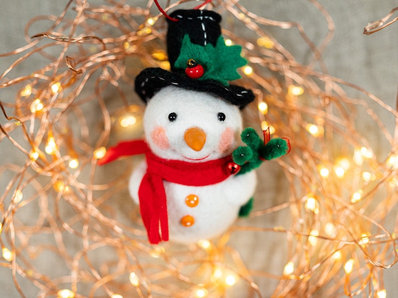 May include: A felt snowman ornament wearing a black top hat with green holly leaves and a red berry, a bright red scarf, and two orange buttons. The snowman has a carrot-shaped nose, pink cheeks, and holds a small green branch with red berries.