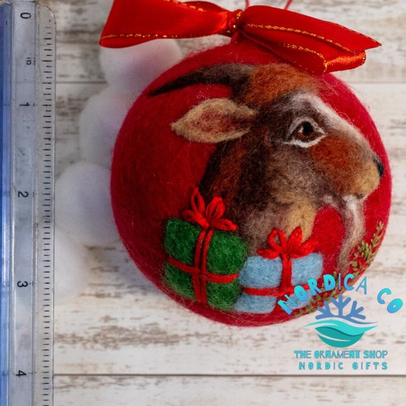 May include: A red felted ball ornament featuring a needle-felted goat head, a green gift box, and a light blue gift box with red ribbons. A red satin ribbon bow sits on top. A clear ruler shows the ornament is approximately 3.5 inches tall.