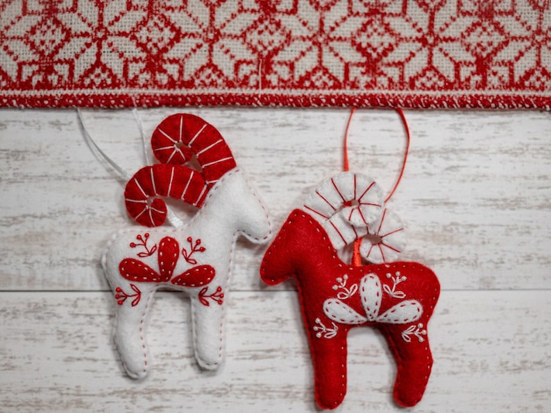 May include: Two felt Christmas ornaments shaped like reindeer with red and white details. The reindeer are hanging from a white string against a white wooden background.