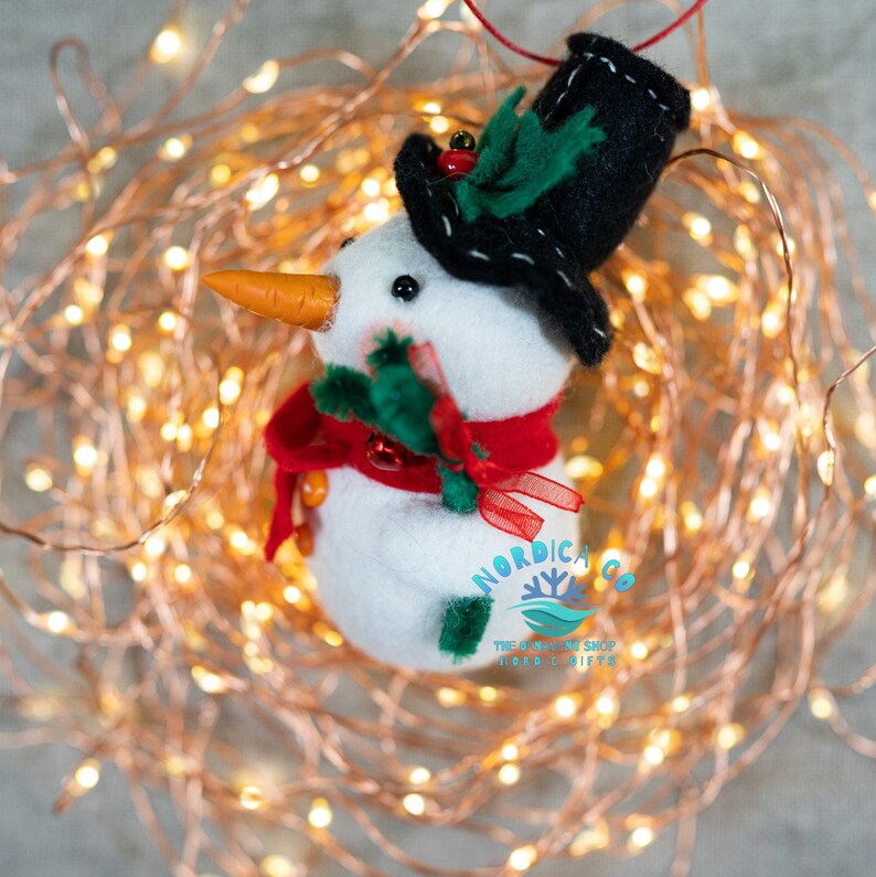 May include: A white felt snowman ornament wearing a black top hat with green holly and a red berry, a red scarf, and an orange carrot nose. The snowman has small black eyes and green felt arms. The text NORDICA CO THE O-MAKING SHOP NORDICA GIFTS is visible.