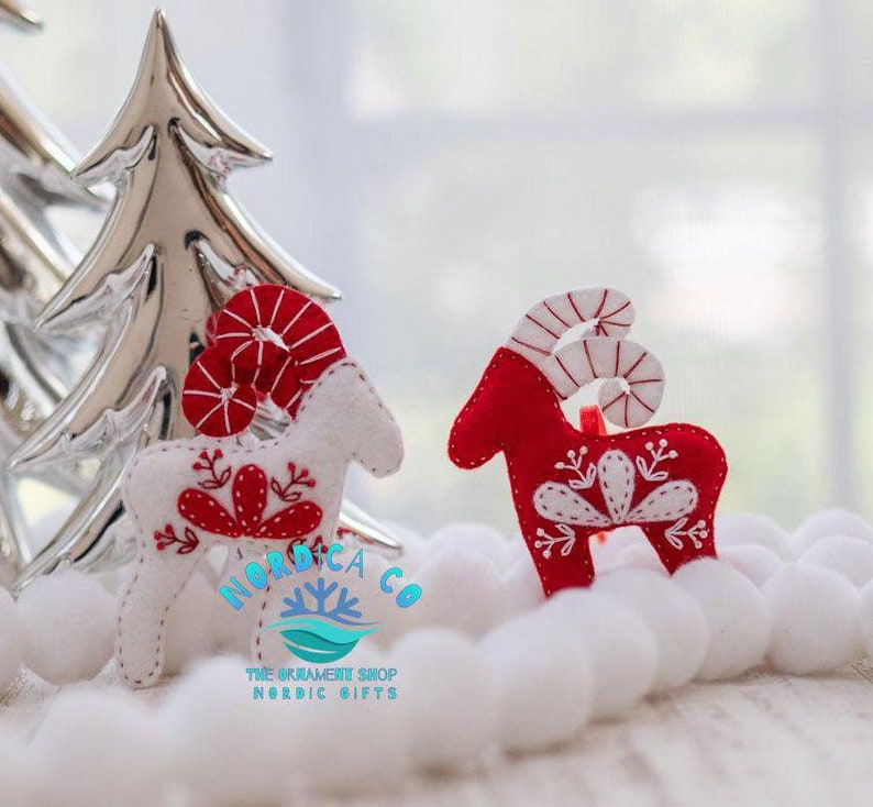 May include: Two felt Christmas ornaments shaped like reindeer with red and white candy cane horns. The reindeer are standing on a white surface. The reindeer on the left is white with red details and the reindeer on the right is red with white details. The ornaments are handmade and have a Scandinavian design.