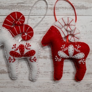 May include: Two felt Christmas ornaments shaped like reindeer with red and white candy cane horns and white embroidered details on a white wood background.