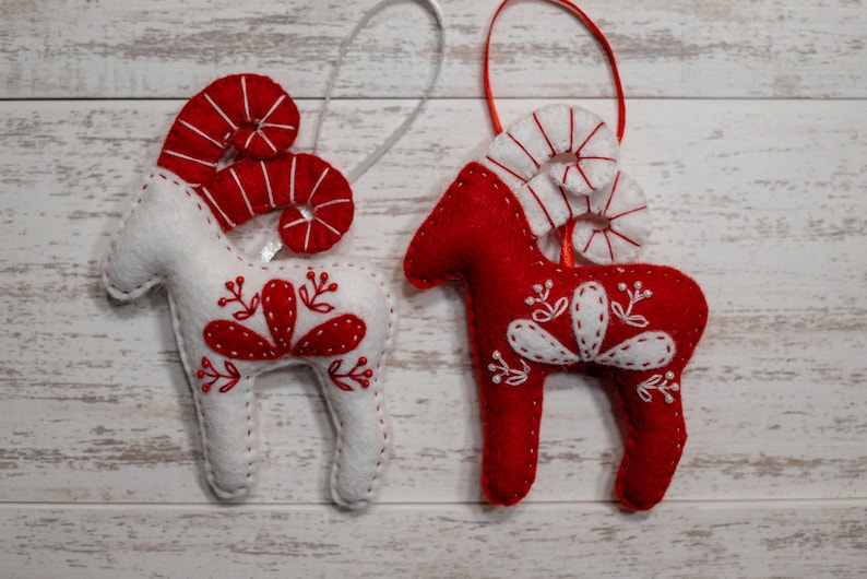May include: Two felt Christmas ornaments shaped like reindeer with red and white candy cane horns and white embroidered details on a white wood background.