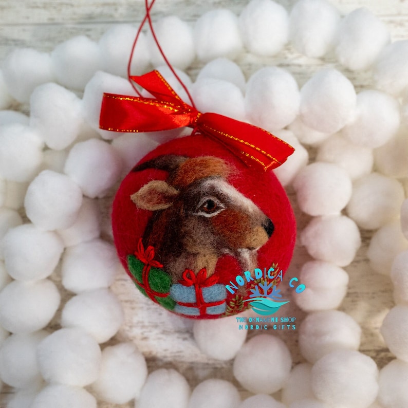 May include: A round red felted ornament featuring a needle-felted brown goat head with a white stripe, positioned above a green and a light blue gift box. A bright red satin ribbon bow is attached to the top with a thin red hanging string.