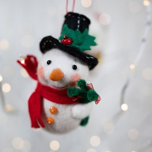 May include: A felt snowman ornament featuring a black top hat with a green holly leaf and red berry accent. The snowman has a bright orange carrot nose, rosy pink cheeks, two orange buttons on its chest, and a red scarf while holding a green branch.