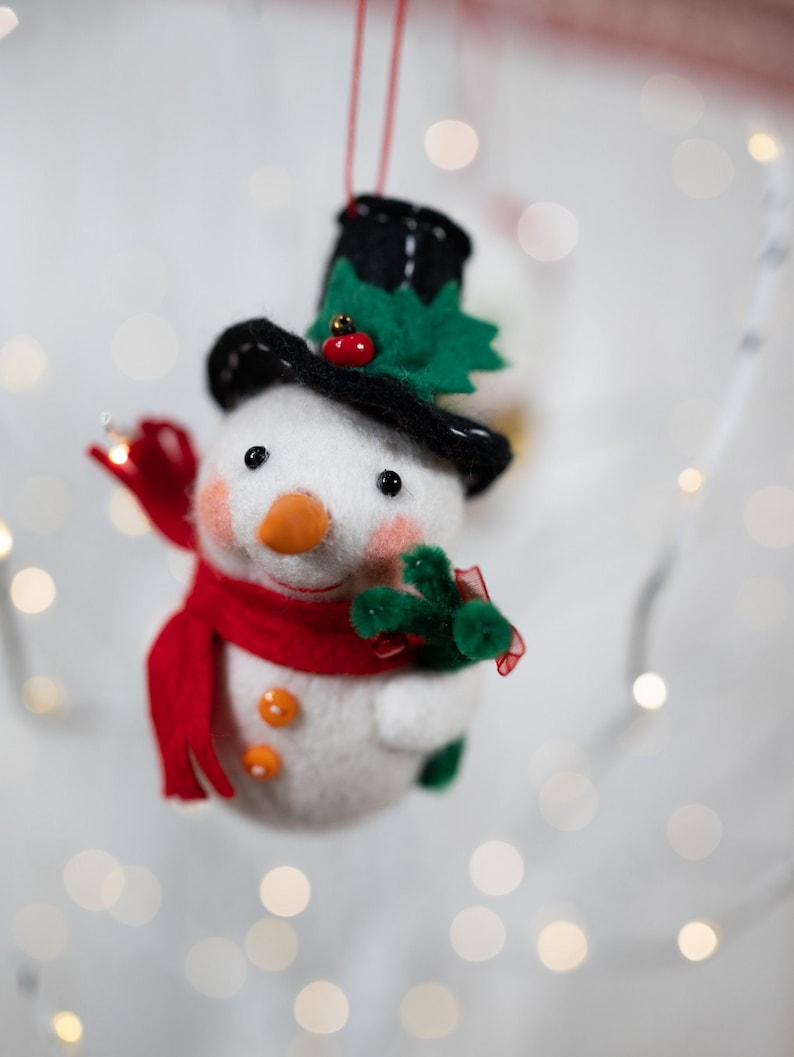 May include: A felt snowman ornament featuring a black top hat with a green holly leaf and red berry accent. The snowman has a bright orange carrot nose, rosy pink cheeks, two orange buttons on its chest, and a red scarf while holding a green branch.