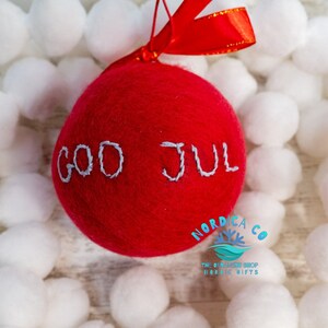 May include: A round, red felted wool ornament featuring the hand-stitched light gray text "GOD JUL" across the center. The ornament is topped with a bright red satin ribbon for hanging, providing a soft, textured appearance for holiday decor.