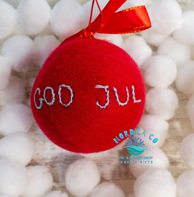 May include: A round, red felted wool ornament featuring the hand-stitched light gray text "GOD JUL" across the center. The ornament is topped with a bright red satin ribbon for hanging, providing a soft, textured appearance for holiday decor.