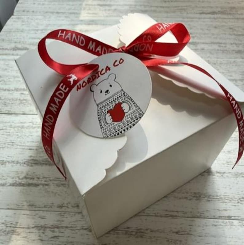 May include: A square white cardboard box tied with a red satin ribbon printed with the words HAND MADE. A round white tag is attached to the ribbon, featuring a black line drawing of a bear wearing a patterned sweater and holding a red heart.