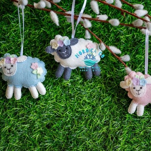 May include: Three felt sheep ornaments with floral headbands hanging from a branch. The sheep are blue, white, and pink.