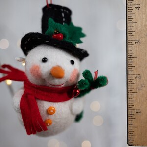 May include: A felt snowman ornament with a black top hat, a red scarf, and two orange buttons. The snowman has a carrot-colored nose, pink cheeks, and green holly leaves with a red berry on the hat. A wooden ruler shows it is 3 inches tall.