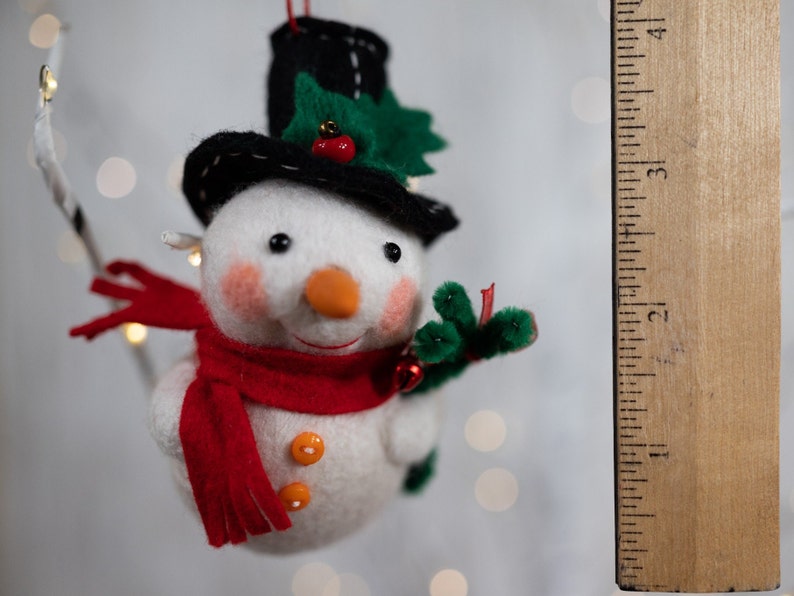 May include: A felt snowman ornament with a black top hat, a red scarf, and two orange buttons. The snowman has a carrot-colored nose, pink cheeks, and green holly leaves with a red berry on the hat. A wooden ruler shows it is 3 inches tall.