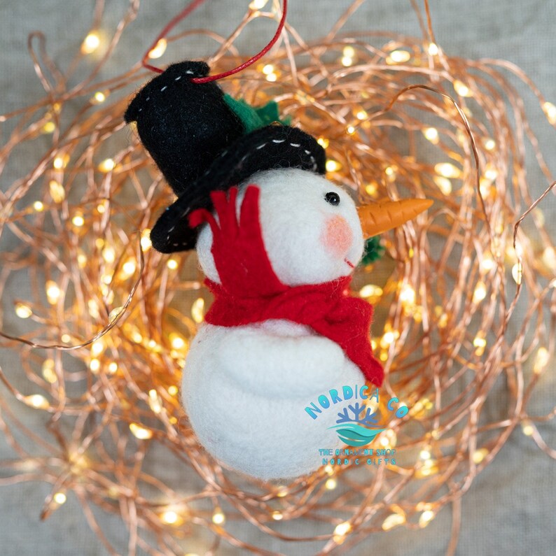 May include: A white felt snowman ornament wearing a black top hat with white stitching and a red scarf. The snowman has a small orange carrot nose, black bead eyes, and pink cheeks. A red string is attached to the top. Text reads NORDICA CO.