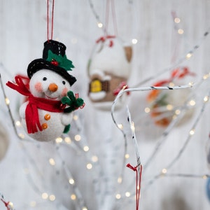 May include: A felt snowman ornament wearing a black top hat with green holly leaves and a red berry, a bright red scarf, and orange buttons. The snowman has a small orange carrot nose and rosy cheeks, hanging from a red string among other ornaments.
