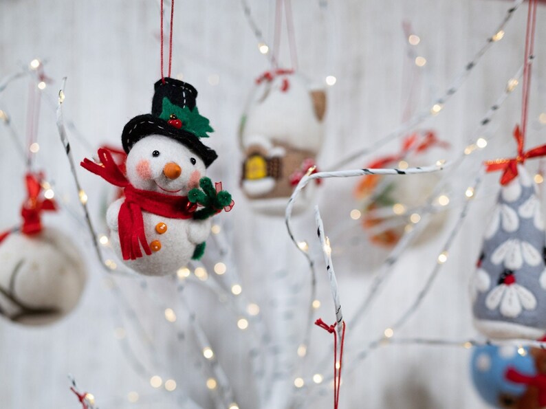 May include: A felt snowman ornament wearing a black top hat with green holly leaves and a red berry, a bright red scarf, and orange buttons. The snowman has a small orange carrot nose and rosy cheeks, hanging from a red string among other ornaments.