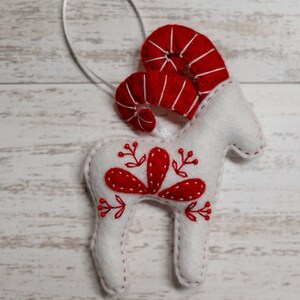 May include: White felt ornament shaped like a ram with red felt horns and red embroidered floral details.