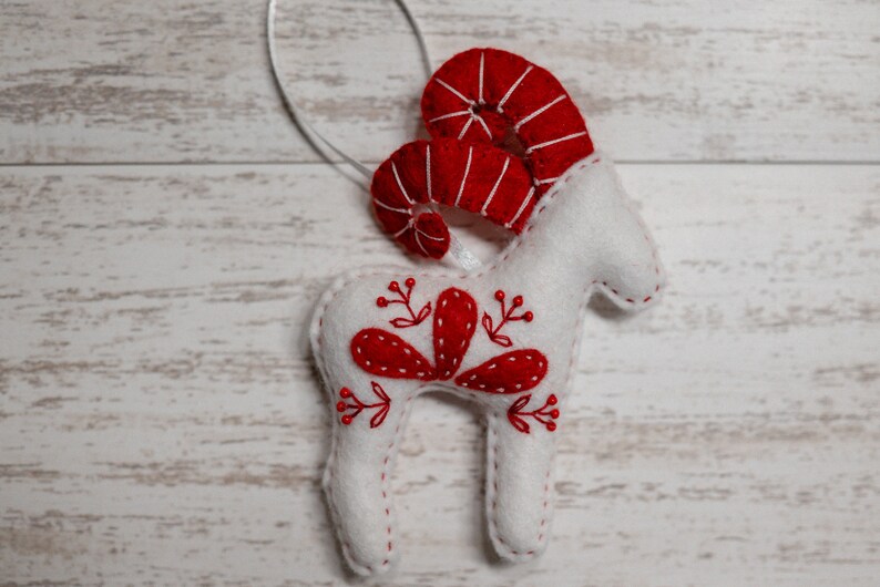 May include: White felt ornament shaped like a ram with red felt horns and red embroidered floral details.