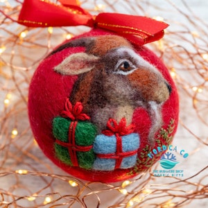 May include: A red felted ball ornament featuring a needle-felted goat head with brown and white fur, positioned above a green gift box and a light blue gift box, both tied with red thread. A red ribbon bow is attached to the top of the ornament.