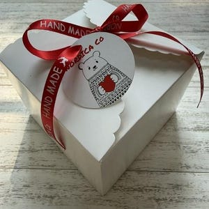 May include: A square white cardboard gift box with scalloped edges, tied with a red satin ribbon printed with the text HAND MADE. A round white tag is attached, featuring a black line drawing of a bear in a patterned sweater holding a red heart.