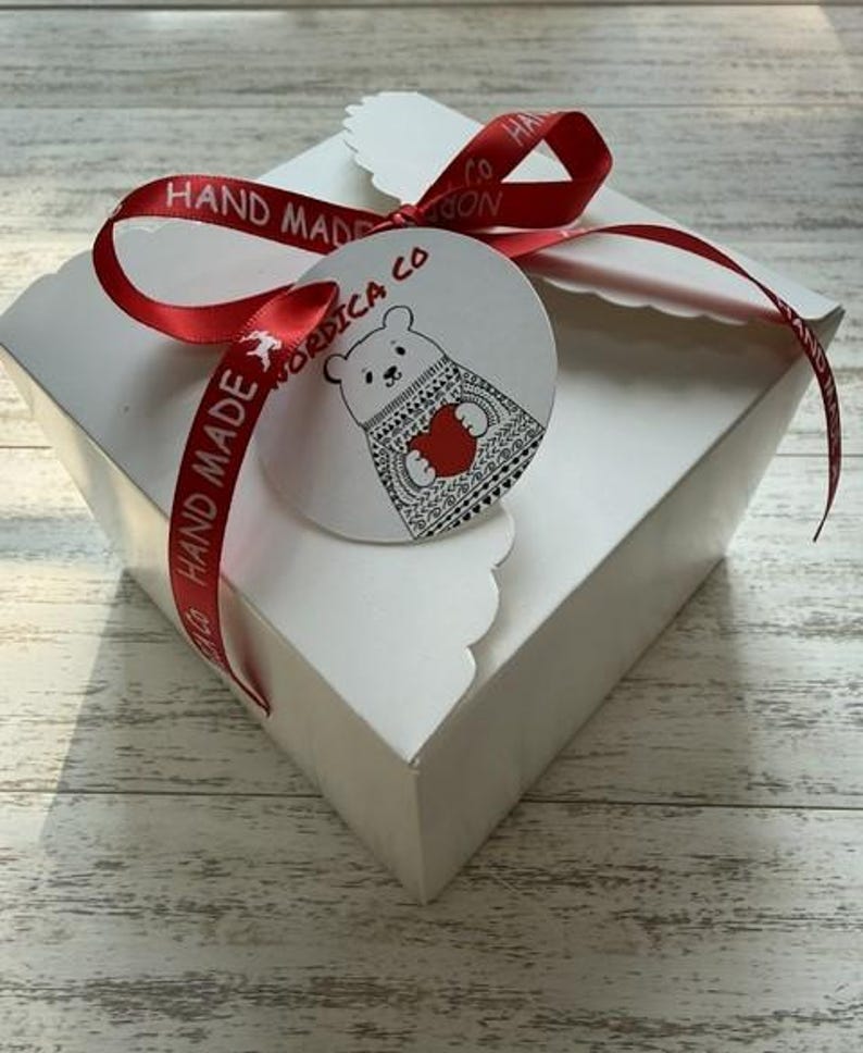 May include: A square white cardboard gift box with scalloped edges, tied with a red satin ribbon printed with the text HAND MADE. A round white tag is attached, featuring a black line drawing of a bear in a patterned sweater holding a red heart.