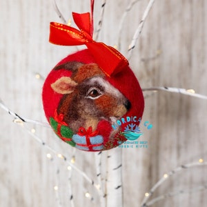 May include: A round red felted Christmas ornament featuring a needle-felted brown goat head with a white stripe, holding two small gift boxes in green and blue. A bright red ribbon bow is attached to the top. The text NORDICA CO is visible.