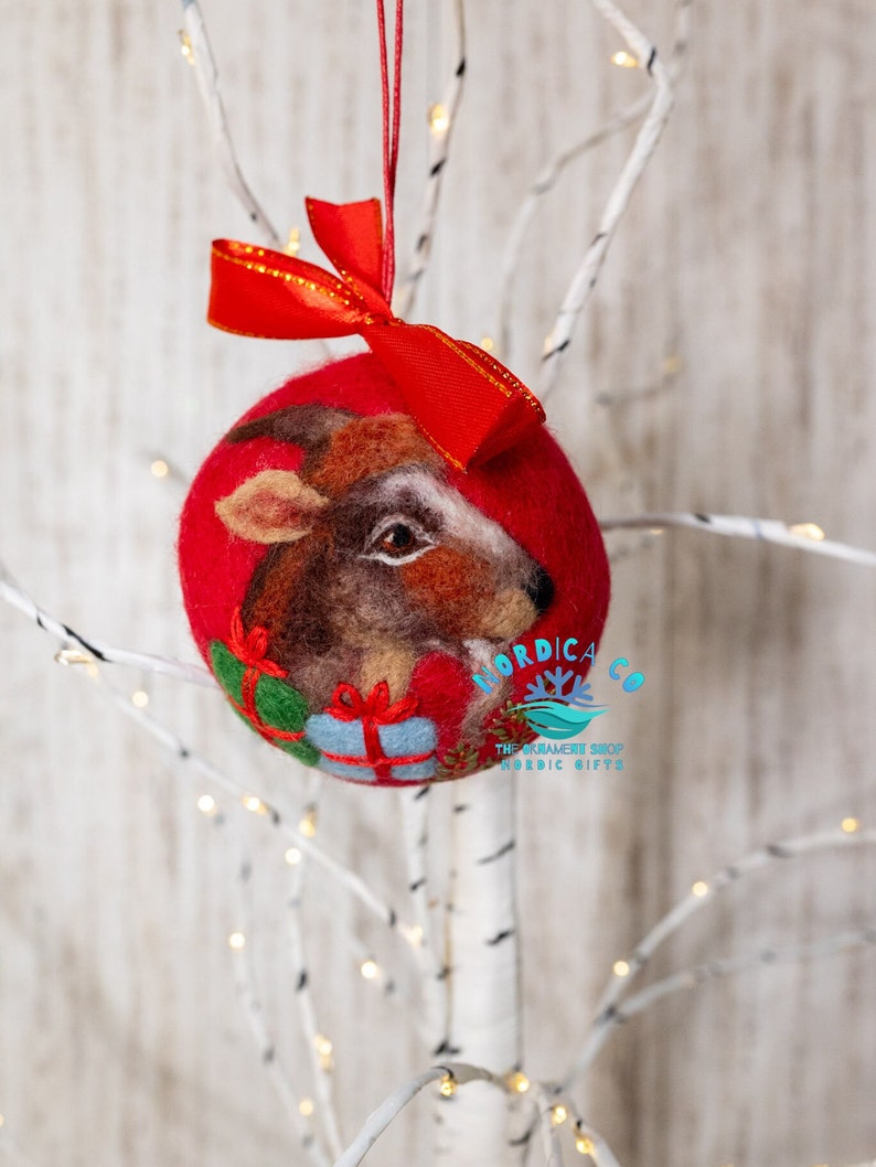 May include: A round red felted Christmas ornament featuring a needle-felted brown goat head with a white stripe, holding two small gift boxes in green and blue. A bright red ribbon bow is attached to the top. The text NORDICA CO is visible.